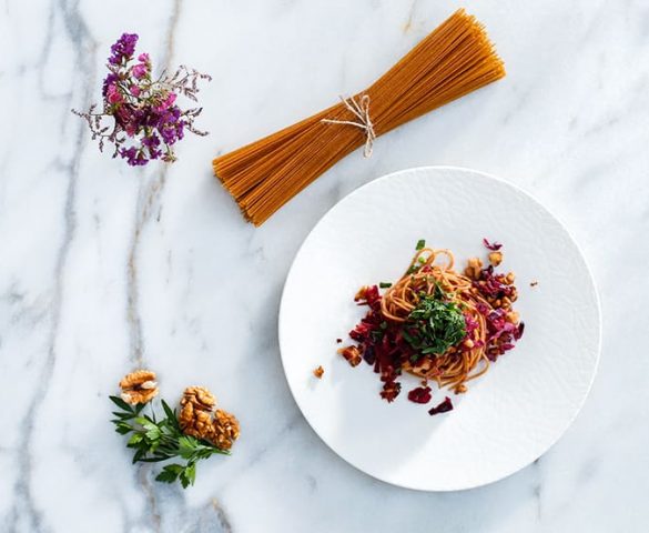 Mermer tezgah üstünde yer alan tabakta cevizli ve kırmızı lahanalı makarna; çevresinde kurutulmuş çiçek, pişirilmemiş spagetti, ayıklanmış ceviz ve bir tutam maydanoz