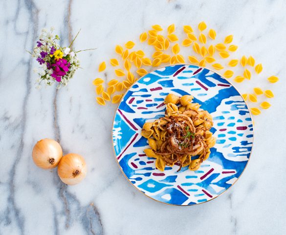 Mermer tezgah üstünde yer alan tabakta soğanlı kremalı makarna; çevresinde kurutulmuş çiçek, pişirilmemiş midye makarna, iki tane kuru soğan