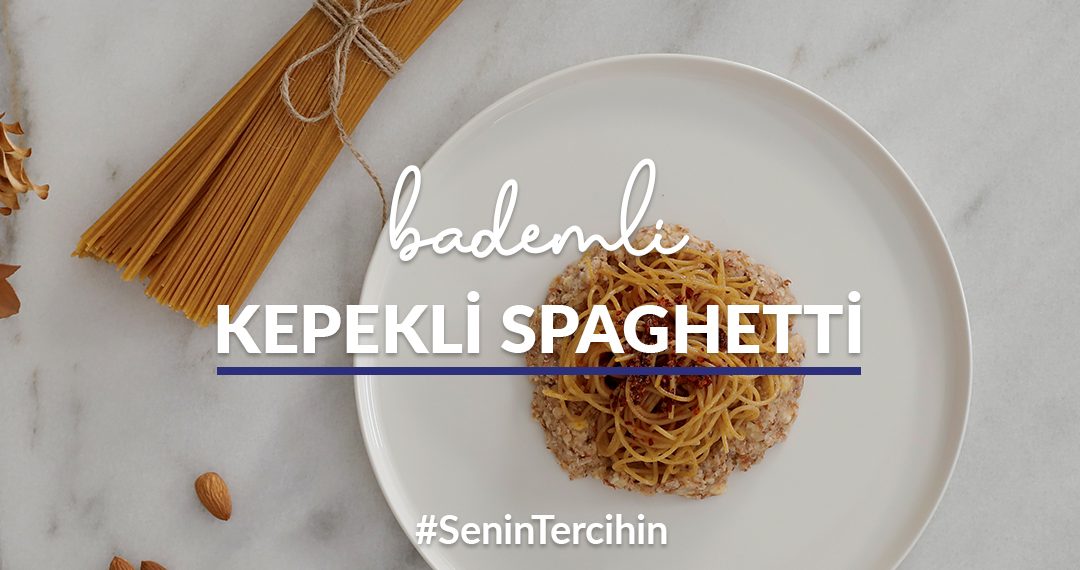 Mermer tezgah üstünde yer alan tabakta bademli kepekli spaghetti; çevresinde taze çiçek, pişirilmemiş kepekli spaghetti ve badem taneleri