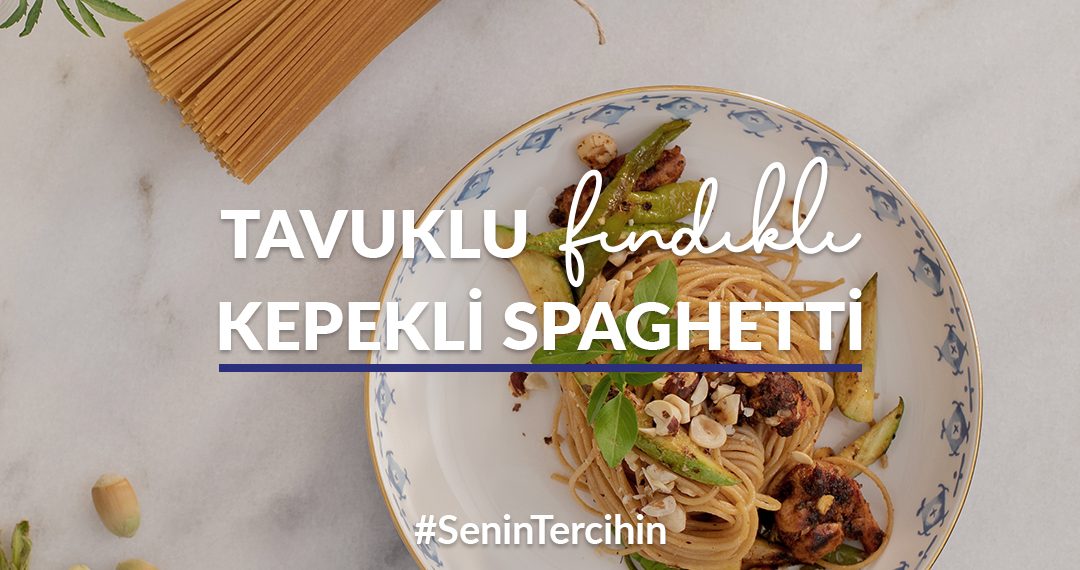 Mermer tezgah üstünde yer alan tabakta tavuklu fındıklı kepekli spaghetti; çevresinde taze çiçek, pişirilmemiş kepekli spaghetti ve dalında koparılmış taze fındık