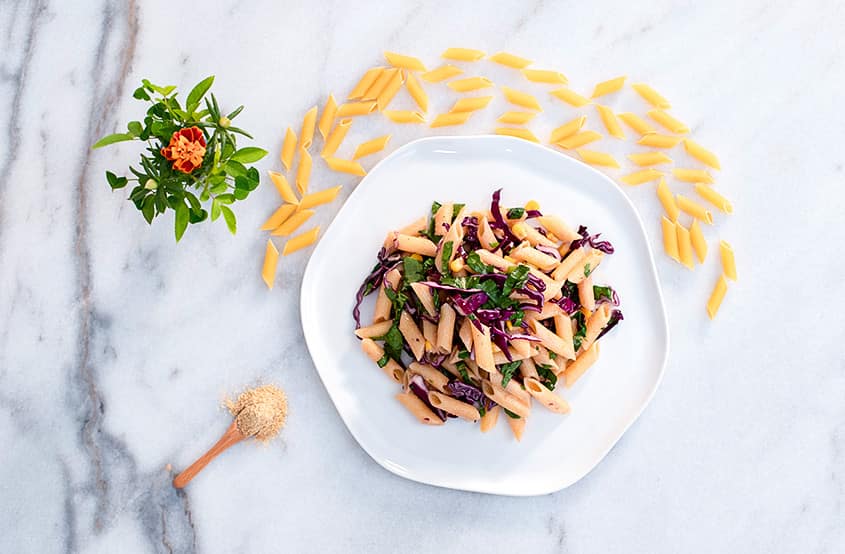 Mermer tezgah üstünde yer alan tabakta hardal soslu makarna salatası; çevresinde taze yeşil ve sarı çiçek, pişirilmemiş penne makarna, tahta kaşıkta hardal tozu